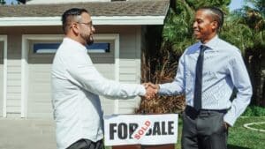 Two men shaking hands in front of house sold sign, sealing real estate deal outdoors.