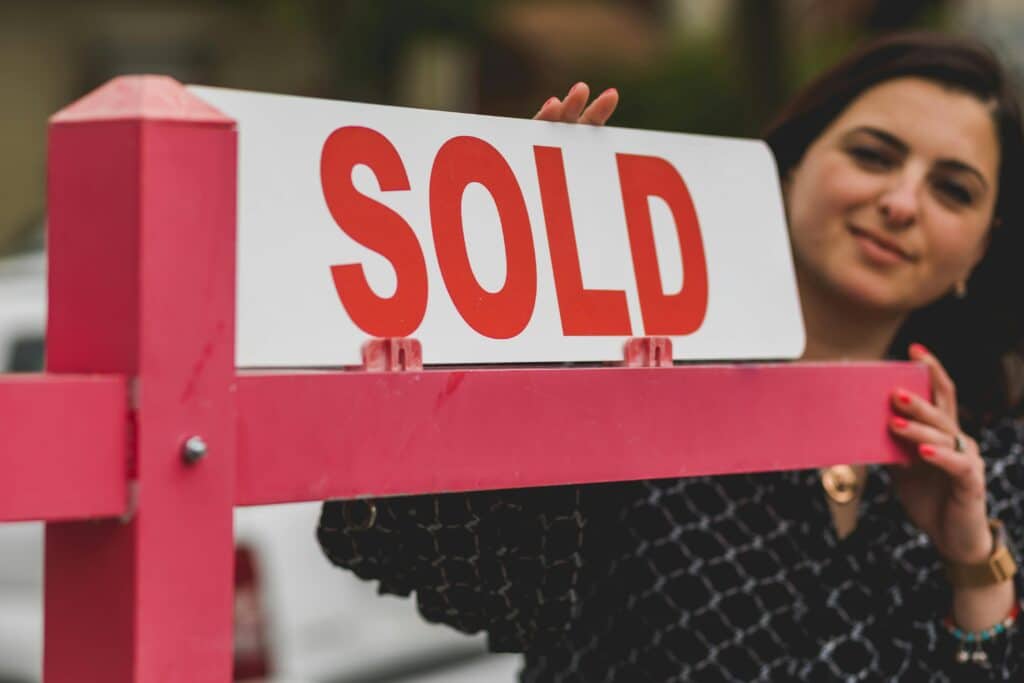 Woman smiling behind a 'Sold' sign, symbolizing successful home purchase.