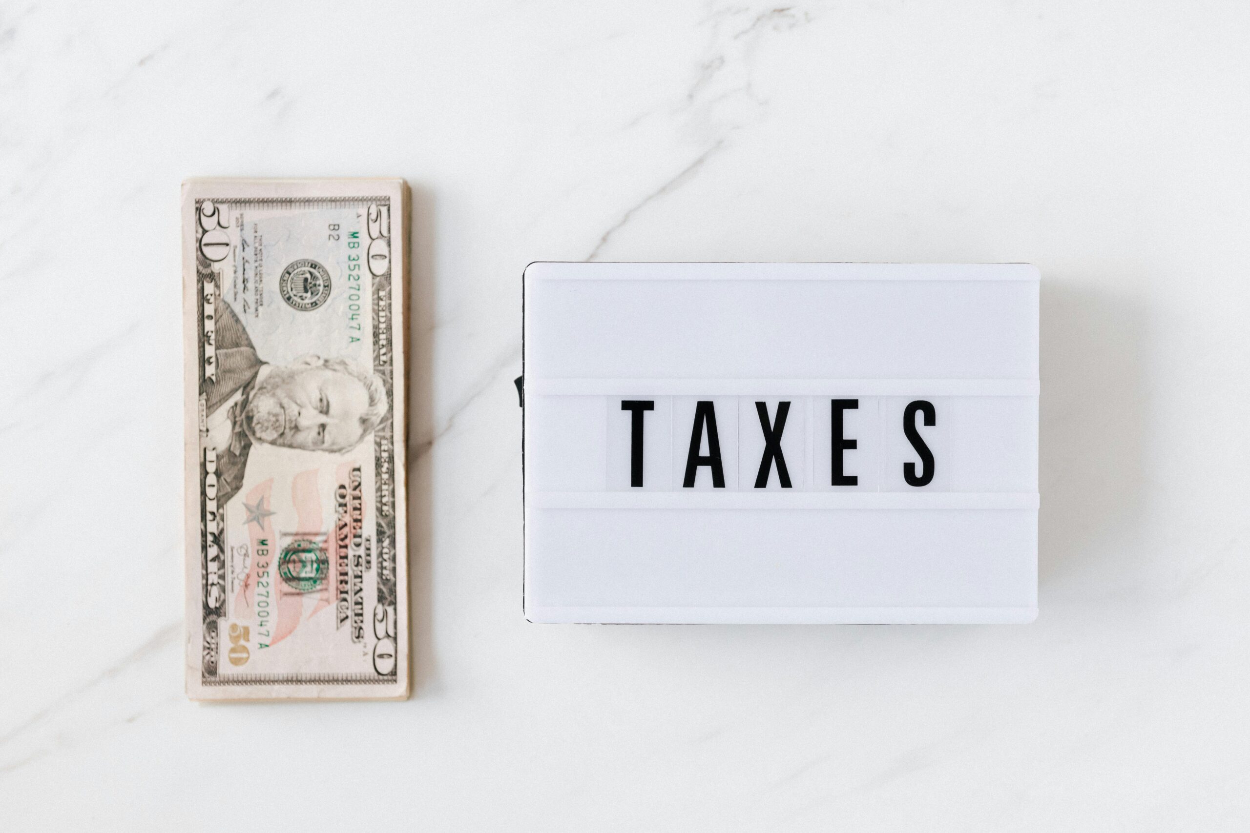 Overhead view of a fifty-dollar bill and a lightbox with 'TAXES' on a marble surface.